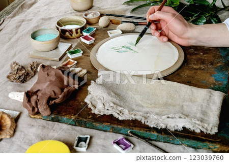 Workspace in ceramic studio, paints and brushes for underglaze painting. Young man's hands painting white plate. Branches, natural decor, linen tablecloth, daylight. Workshop idea. Workspace in ceramic studio, paints and brushes for underglaze painting. Young man's hands painting white plate. Branches, natural decor, linen tablecloth, daylight. Workshop idea. 123039760