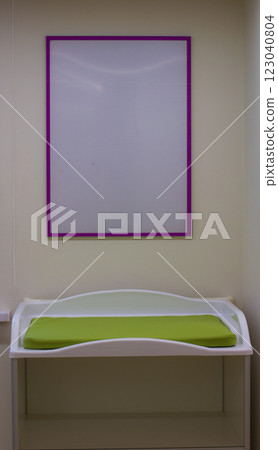 White baby changing table with green mattress and empty purple frame hanging on the wall in a nursery, representing childcare and baby care facilities. White baby changing table with green mattress and empty purple frame hanging on the wall in a nursery, representing childcare and baby care facilities. 123040804