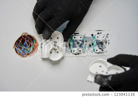 Close-up of an electrician wearing black gloves installing power socket on white wall. Electrical repair and maintenance process in progress 123041138