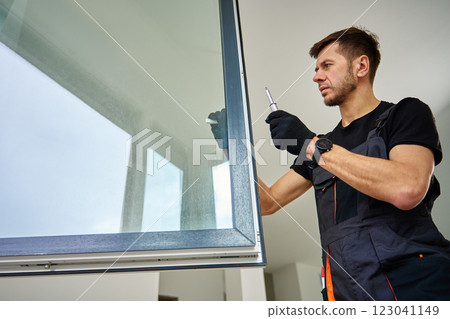 Skilled worker wearing gloves uses screwdriver to fix window frame, home renovation tasks. Professional repairman adjusting white pvc windows in modern building Skilled worker wearing gloves uses screwdriver to fix window frame, home renovation tasks. Professional repairman adjusting white pvc windows in modern building 123041149