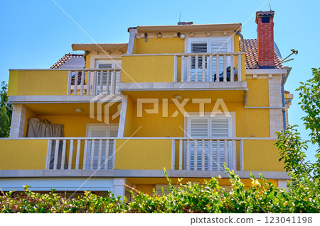 Vibrant yellow villa with white shutters, balcony and lush greenery. Traditional architecture in Croatian village. Rental house for summer vacations in coastal town 123041198