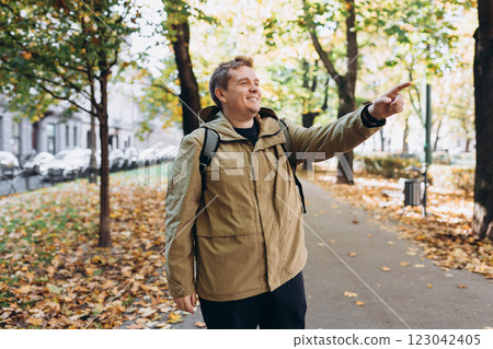 Blonde young 30s man wearing backpack pointing something on city autumn park street. Urban lifestyle, travel concept. High quality photo 123042405