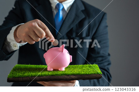 Businessman puts coin in piggy bank standing on grass. Concept of saving and investment and savings. Personal finance, budgeting. Financial growth. Wealth accumulation, smart money management. 123043316