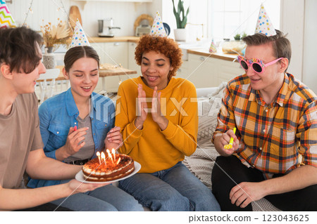 Make a wish. Woman wearing party cap blowing out burning candles on birthday cake. Happy Birthday party. Group of friends wishes girl happy birthday. People celebrating birthday with party at home Make a wish. Woman wearing party cap blowing out burning candles on birthday cake. Happy Birthday party. Group of friends wishes girl happy birthday. People celebrating birthday with party at home 123043625