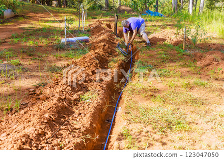 Plumber works diligently to dig trench install an PVC water pipe irrigation line in lush garden surrounded by trees. 123047050