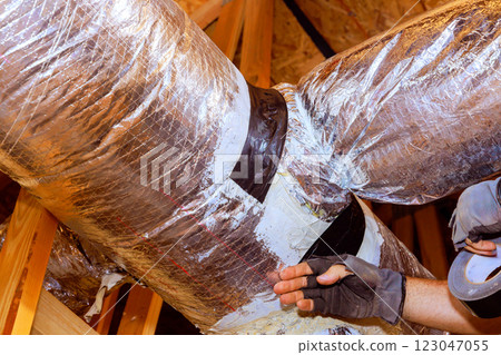 Skilled technician repairs ductwork, insulation while working in residential attic during works Skilled technician repairs ductwork, insulation while working in residential attic during works 123047055