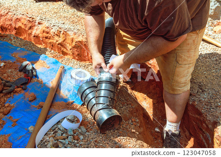 Skilled worker connects drainage pipe while kneeling in trench at construction site under works 123047058