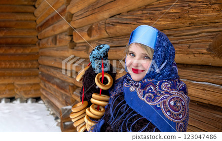 Woman in Russian folk headdress with bunch of bagels on Shrovetide near wooden building made of logs 123047486