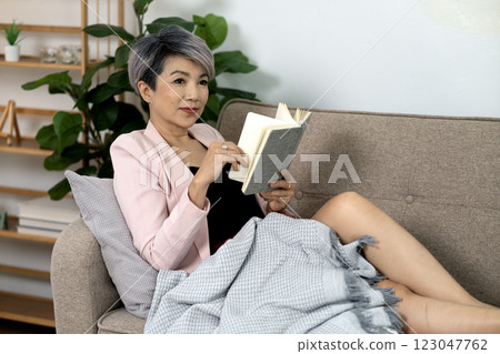 Senior woman in business look is sitting on sofa relaxing reading book 123047762