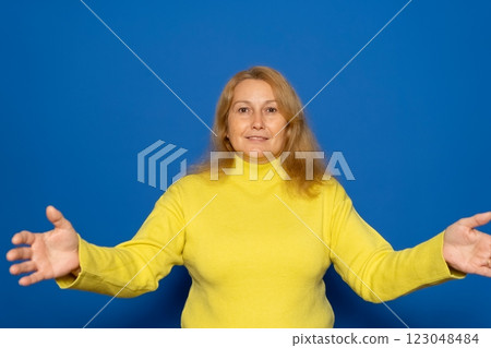 Caucasian woman in her 40s wearing a yellow sweater with her arms open in a welcoming gesture, isolated on blue studio background. 123048484