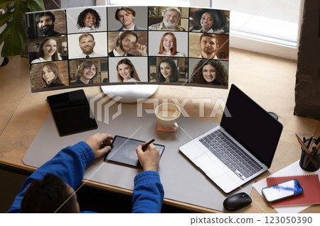 Remote worker using tablet and pen while engaged in video meeting with team displayed on monitor against organized home office. 123050390