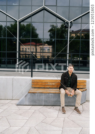 Full length vertical portrait of thoughtful man sitting on modern bench in city street, holding two disposable coffee cups, enjoying break in front of modern building. Concept of urban lifestyle. Full length vertical portrait of thoughtful man sitting on modern bench in city street, holding two disposable coffee cups, enjoying break in front of modern building. Concept of urban lifestyle. 123051603