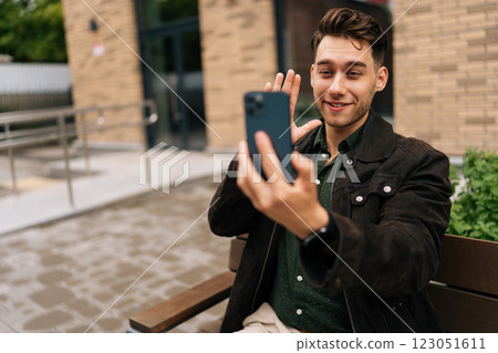 Portrait of smiling young man sitting on urban bench, waving hand while engaging in video call on smartphone in modern city environment. Concept of casual communication and connectivity. Portrait of smiling young man sitting on urban bench, waving hand while engaging in video call on smartphone in modern city environment. Concept of casual communication and connectivity. 123051611