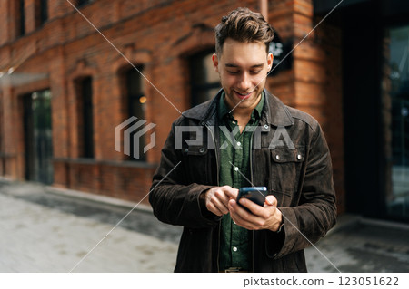 Medium shot portrait of stylish young man in suede jacket using smartphone standing on modern city street with red brick buildings on summer day. Concept of modern communication, urban lifestyle. 123051622