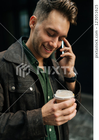 Close-up vertical portrait of handsome smiling man in casual attire, holding takeaway coffee cup, having phone conversation standing on urban street. Concept of successful city lifestyle 123051625