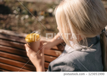 A blonde woman sits on a bench holding a cup of coffee. The bench is wooden and the woman is wearing a gray jacket. 123051990
