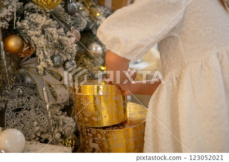 A woman is opening a gold box with a silver ball inside. The box is decorated with gold and silver ornaments, and the woman is wearing a white dress. The scene is set in a room with a Christmas tree. 123052021
