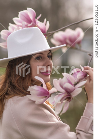 Woman magnolia flowers, surrounded by blossoming trees., hair down, white hat, wearing a light coat. Captured during spring, showcasing natural beauty and seasonal change. Woman magnolia flowers, surrounded by blossoming trees., hair down, white hat, wearing a light coat. Captured during spring, showcasing natural beauty and seasonal change. 123052110