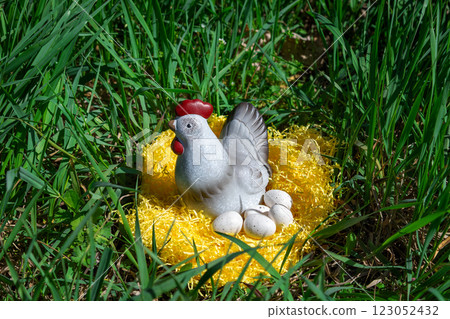 Decorative ceramic hen with speckled eggs on yellow straw in green grass. Spring and Easter symbol, representing fertility, nature, and seasonal celebration. Decorative ceramic hen with speckled eggs on yellow straw in green grass. Spring and Easter symbol, representing fertility, nature, and seasonal celebration. 123052432