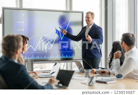 A businessman stands at the front of a meeting room, pointing at a large screen displaying a financial graph. He is presenting data to his colleagues, who are seated around a conference table. A businessman stands at the front of a meeting room, pointing at a large screen displaying a financial graph. He is presenting data to his colleagues, who are seated around a conference table. 123053087