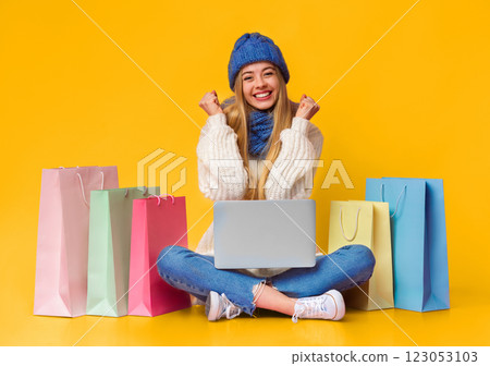 Finally big discounts. Girl in knitted hat and scarf clenching fists and smiling, sitting on floor with laptop among shopping bags, copy space 123053103