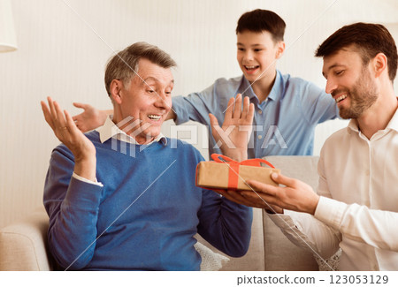Middle-Aged Son And Grandson Congratulating Elderly Man Giving Him Birthday Gift Sitting On Couch Indoor. Selective Focus 123053129