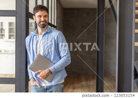 A young man with a beard is leaning against a glass and metal doorway while holding a closed laptop computer. He is dressed in a blue button-down shirt over a white t-shirt and blue jeans, copy space A young man with a beard is leaning against a glass and metal doorway while holding a closed laptop computer. He is dressed in a blue button-down shirt over a white t-shirt and blue jeans, copy space 123053154