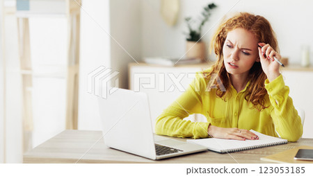 A young woman with red hair sits at a table with a laptop, notepad, and pen, looking thoughtful and working. 123053185