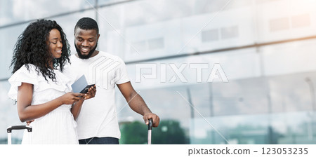 African American couple stands in front of a large glass window at an airport terminal, checking their boarding pass. They appear to be ready to travel and are smiling, copy space 123053235