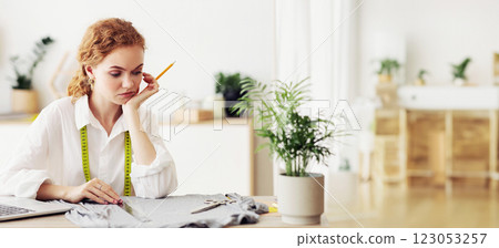 A woman with red hair and a white shirt sits at a desk, measuring fabric with a tape measure. A woman with red hair and a white shirt sits at a desk, measuring fabric with a tape measure. 123053257