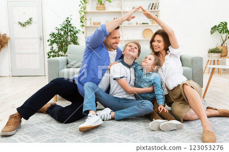 Housing for family. Young parents joining hands making symbolic house roof sitting with children on floor in new apartment. 123053276