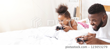African American father and his daughter lie in bed together, both engrossed in playing video games with handheld controllers. They appear to be having a lot of fun together 123053279