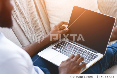 A man is sitting on a couch, working on a laptop. He is wearing blue jeans and a white shirt, and his hands are on the keyboard. The laptop is open and the screen is black, cropped A man is sitting on a couch, working on a laptop. He is wearing blue jeans and a white shirt, and his hands are on the keyboard. The laptop is open and the screen is black, cropped 123053283