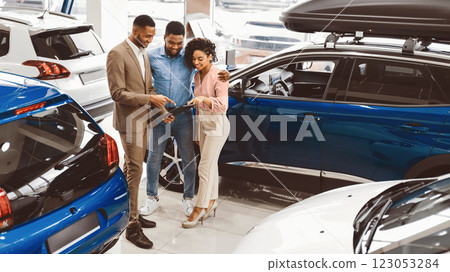 Black Couple Visiting Car Dealership Buying New Vehicle Talking With Salesman Standing In Auto Showroom. Happy Buyers. Panorama, Free Space Black Couple Visiting Car Dealership Buying New Vehicle Talking With Salesman Standing In Auto Showroom. Happy Buyers. Panorama, Free Space 123053284