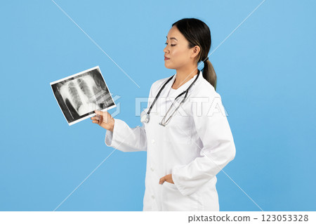 A healthcare worker Asian lady in a white coat analyzes a chest X-ray while standing against a light blue backdrop. The individual appears focused and engaged in their medical examination. 123053328