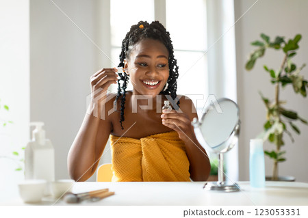 African American young woman with long black braids applies facial serum to her skin, looking in a handheld mirror while seated at a vanity table. She is smiling brightly African American young woman with long black braids applies facial serum to her skin, looking in a handheld mirror while seated at a vanity table. She is smiling brightly 123053331