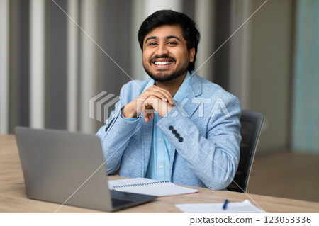 A young Indian man in a light blue blazer sits at a desk in a modern office. He is looking directly at the camera and smiling. His hands are clasped in front of him, and he has a laptop open 123053336