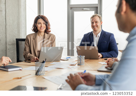 Three business professionals are sitting around a table in a modern office, using laptops and working on projects. Papers and writing utensils are spread out on the table 123053352