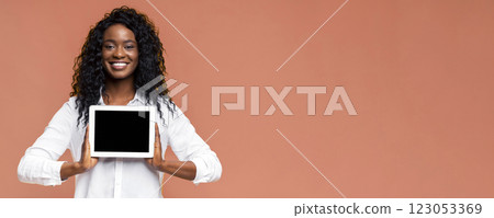 Black woman with curly black hair is smiling and holding a tablet with a blank screen in front of a peach colored background. She is wearing a white button-up shirt. 123053369