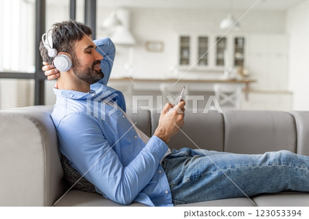 A man is sitting on a couch, wearing headphones and using his smartphone. He appears to be relaxed and enjoying his time. The man is wearing a light blue shirt and blue jeans A man is sitting on a couch, wearing headphones and using his smartphone. He appears to be relaxed and enjoying his time. The man is wearing a light blue shirt and blue jeans 123053394