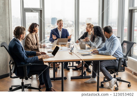 A group of five professionals are seated around a large, rectangular table in a modern office. They are engaged in a discussion, looking at each other and using laptops 123053402
