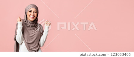 Excited saudi girl in headscarf celebrating success, raising hands and clenching fists, posing over pink background with empty space 123053405