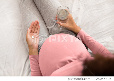Pregnancy medicines. Expectant woman holding pill and glass of water, sitting on bed, top view 123053406
