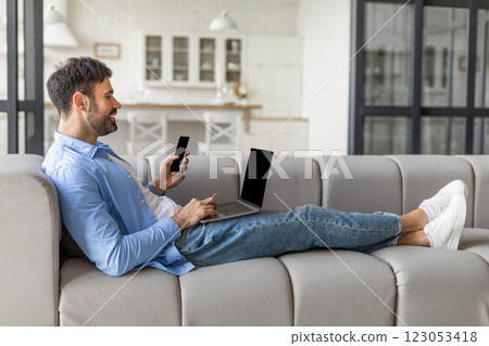 A man is sitting on a gray couch in a modern living room, using his laptop and looking at his smartphone. He is wearing a light blue shirt and blue jeans, and his feet are up on the couch. 123053418
