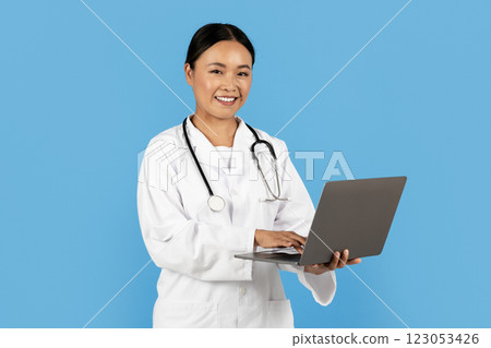 A healthcare professional in a white coat stands with a laptop, smiling while looking directly at the camera. The backdrop is a bright blue, enhancing the inviting atmosphere of the clinic. 123053426