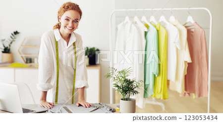 A woman with red hair, wearing a white shirt and a measuring tape, smiles while standing in a studio, with clothes hanging on a rack in the background. 123053434