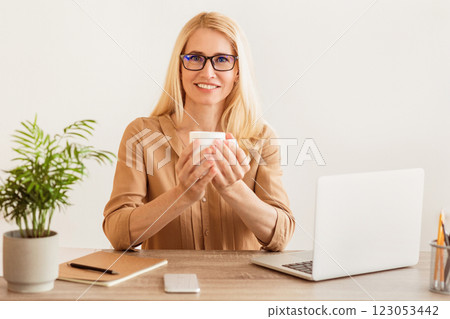 A woman sits at a desk, holding a coffee cup in her hands, with a laptop open in front of her. A woman sits at a desk, holding a coffee cup in her hands, with a laptop open in front of her. 123053442