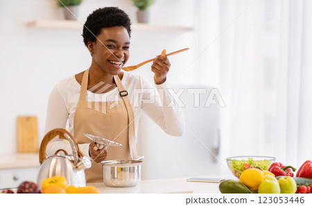 Happy African American Woman Cooking Tasting Dinner In A Pot Standing In Modern Kitchen At Home. Housewife Preparing Healthy Food Smiling To Camera. Household And Nutrition. Dieting Recipes Concept 123053446