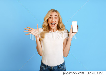 A young woman with long blonde hair is standing in front of a blue background, holding a smartphone with a blank screen. She is wearing a white t-shirt and jeans and has an excited expression A young woman with long blonde hair is standing in front of a blue background, holding a smartphone with a blank screen. She is wearing a white t-shirt and jeans and has an excited expression 123053455