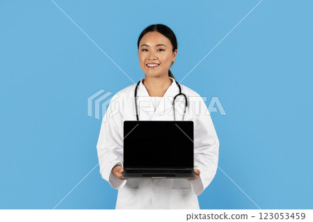 A smiling Asian woman healthcare worker in a white lab coat holds a laptop against a blue backdrop, symbolizing the integration of technology in patient care and medical practice. A smiling Asian woman healthcare worker in a white lab coat holds a laptop against a blue backdrop, symbolizing the integration of technology in patient care and medical practice. 123053459